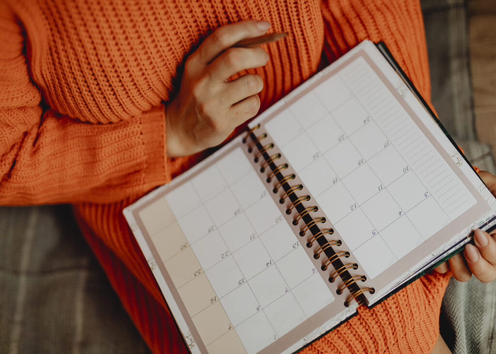 personne-avec-un-pull-orange-qui-tient-un-stylo-dans-la-main-et-un-agenda-pose-sur-ses-genoux