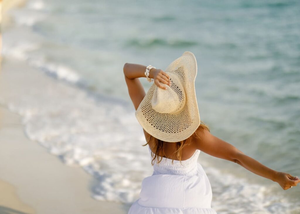 vu-arriere-d-une-femme-en-robe-blanche-en-tenant-son-chapeau-face-a-la-mer